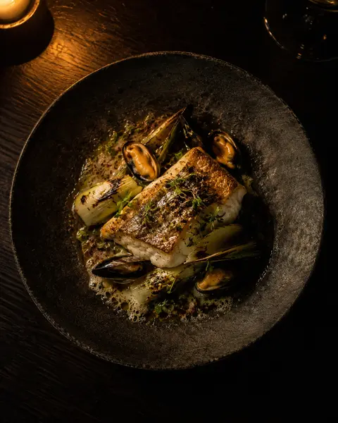 Roast hake with brown butter, charred leeks and mussels on a dark stoneware plate.